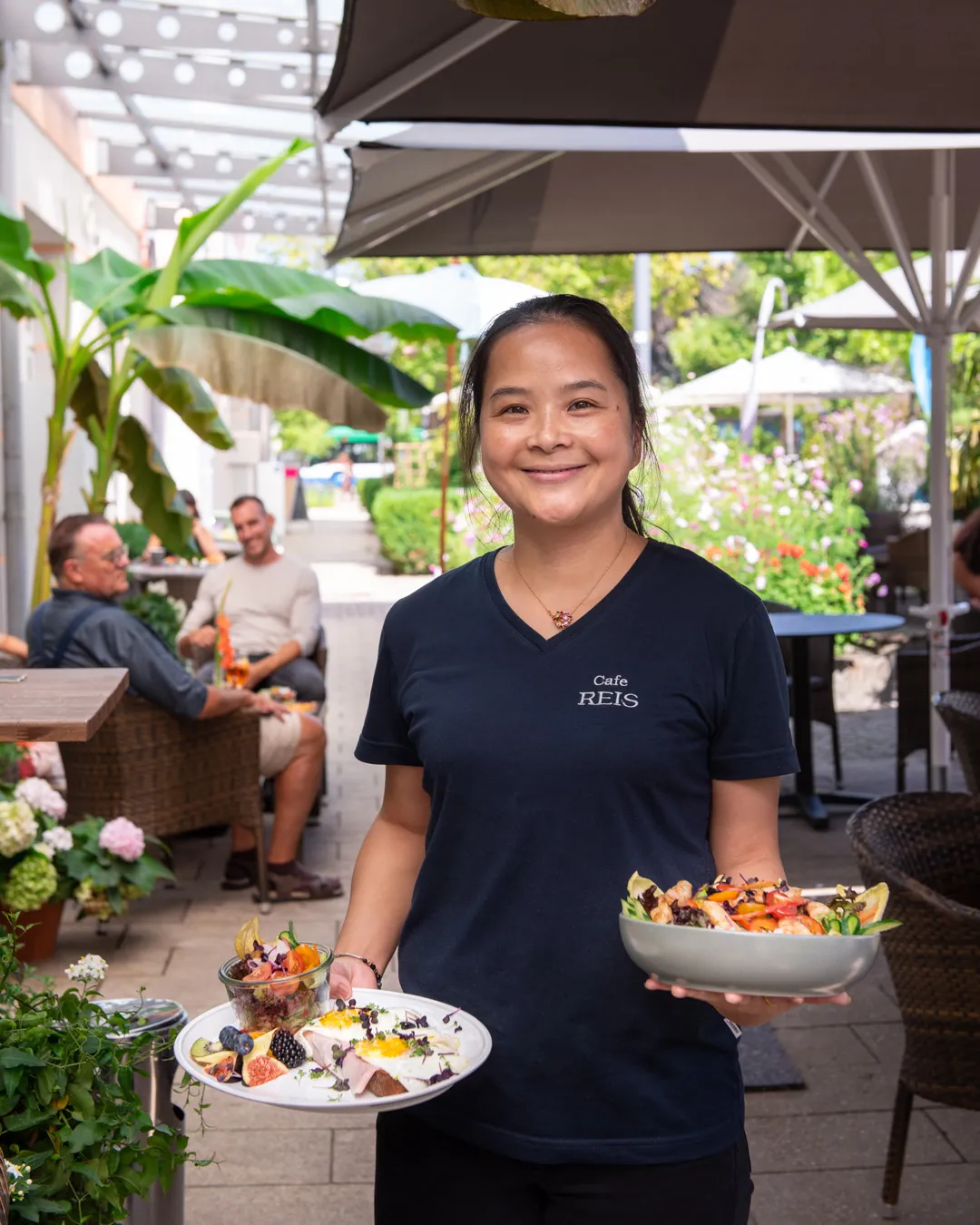 Lächelnde Bedienung im Café Reis Gilching serviert Frühstücksteller und Salatbowl auf der Terrasse unter Palmen