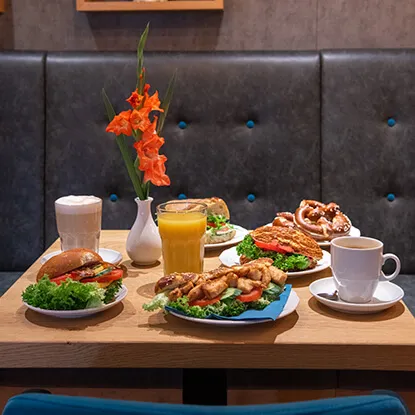 Belegte Baguettes und Bagels mit Salat auf Tisch im Café Reis Gewerbe-Süd Gilching, dazu Cappuccino, Orangensaft und Gladiole