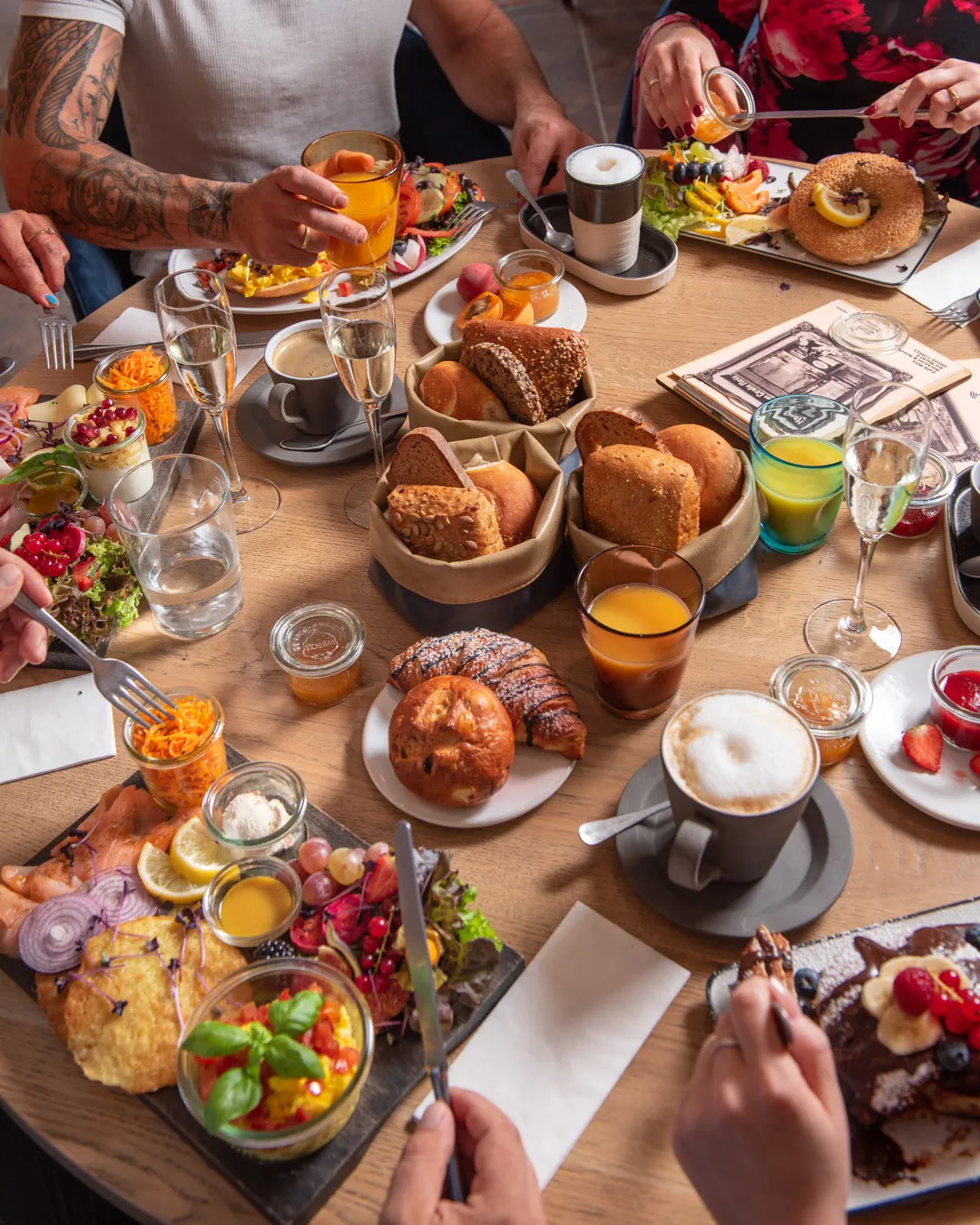 Vier Gäste beim gemeinsamen Brunch im Café Reis Gilching mit Brotkörben, Reiberdatschi, Salat und Prosecco