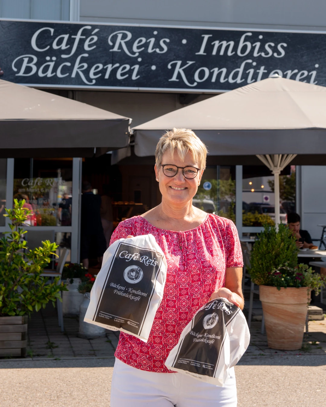 Inhaberin des Café Reis Gilching mit frisch gepackten Bäckerei-Tüten vor dem Laden am Gewerbe-Süd
